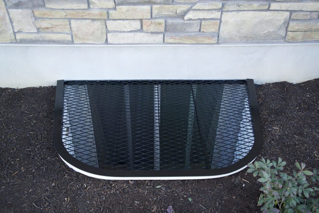 Black metallic window well cover on stone wall with surrounding mulch and plants.