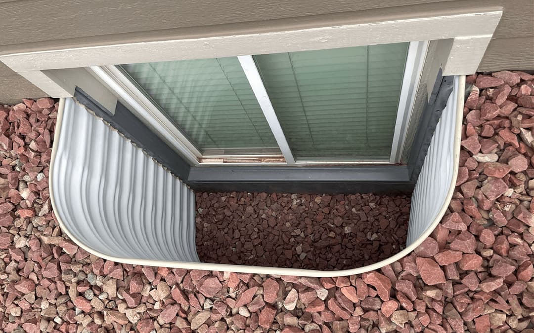 Exterior window well filled with red gravel, featuring a metal liner and nearby window.