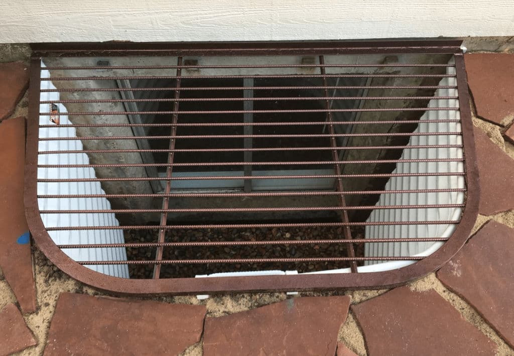 Iron vent grate covering a basement window, surrounded by stone pavement.