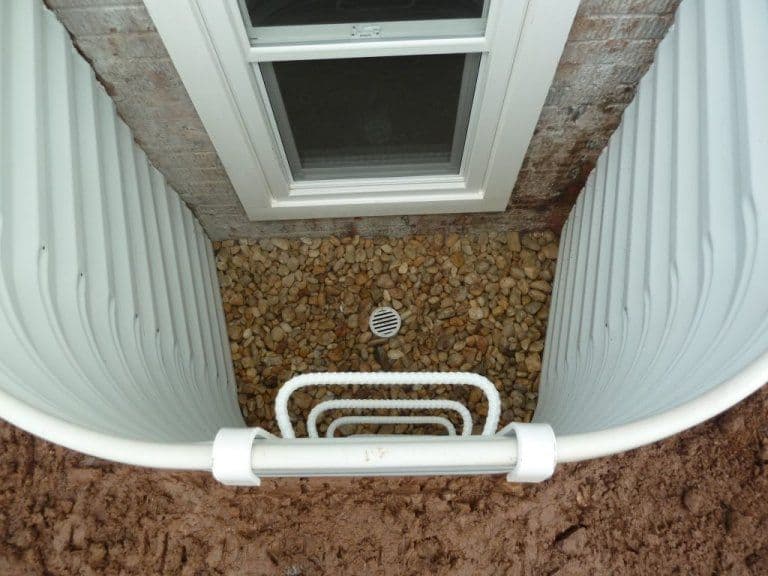 Exterior window well with gravel drainage and white metal frame, installed beside a home.