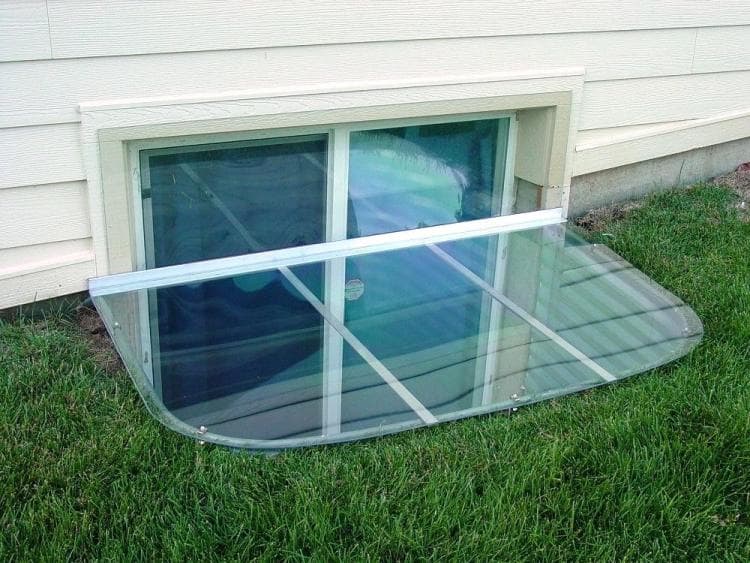 Basement window well with glass cover, showcasing landscaping and sunlight reflection.