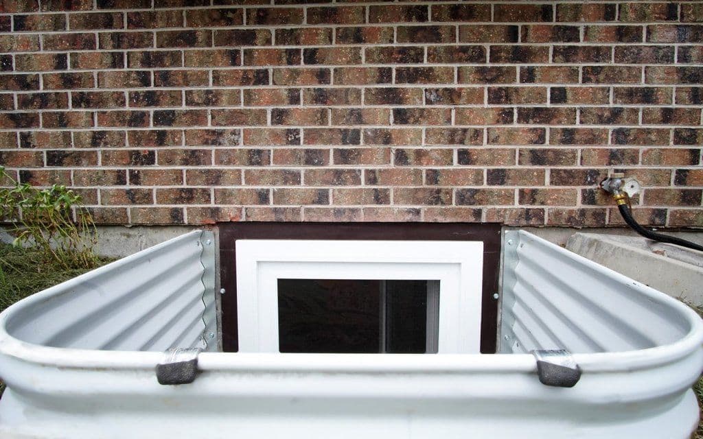 Exterior view of a basement window well with a brick wall background.