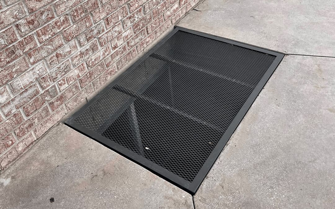 Metal grate covering a basement access point on a concrete sidewalk by a brick wall.