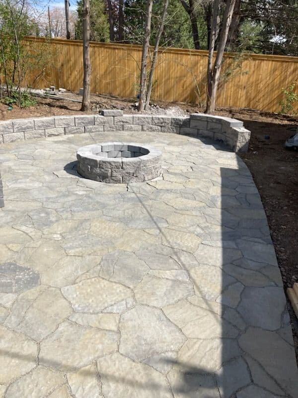 Stone patio with a circular fire pit, surrounded by landscaping and wooden fencing.