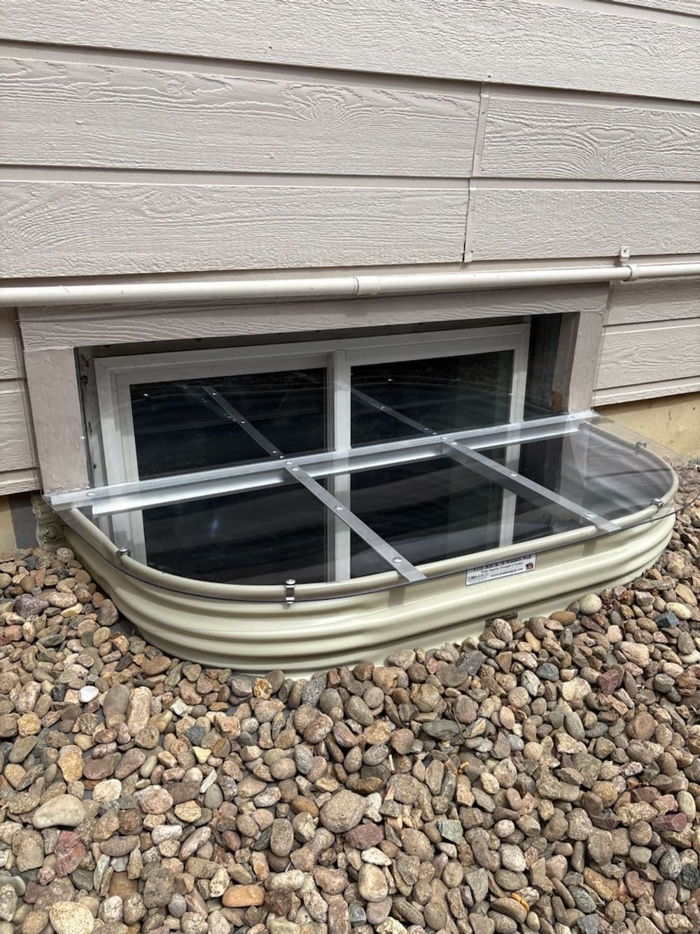 Window well with protective cover installed outside a home, surrounded by decorative gravel.